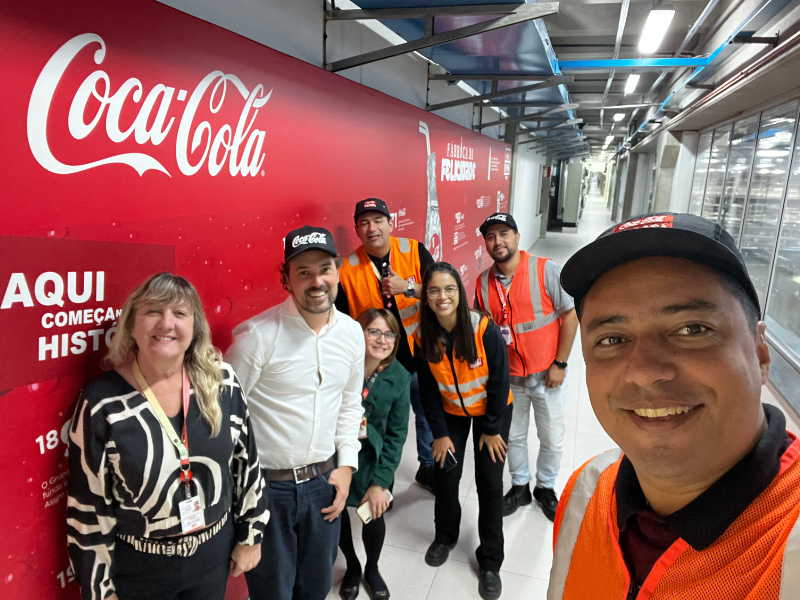 Grupo de pessoas, incluindo Djalma, sócio da Egalite, e outros colaboradores com coletes de segurança, reunidos em frente a um mural da Coca-Cola em uma instalação da empresa, simbolizando parceria e trabalho colaborativo em um ambiente industrial.