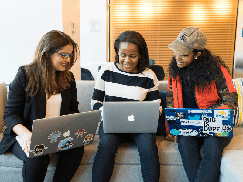 Três mulheres de diferentes origens sentadas em um sofá, colaborando em um projeto usando seus laptops. A imagem transmite diversidade e trabalho em equipe em um ambiente inclusivo e tecnológico.