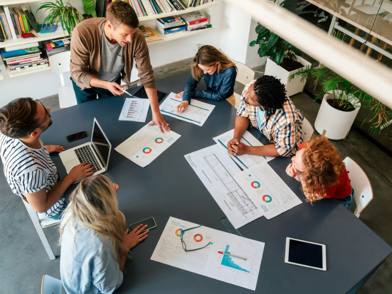 Equipe diversificada reunida em uma sala de reunião, discutindo gráficos e relatórios espalhados sobre uma mesa. A imagem representa colaboração, diversidade e análise de dados em um ambiente de trabalho moderno e inclusivo.