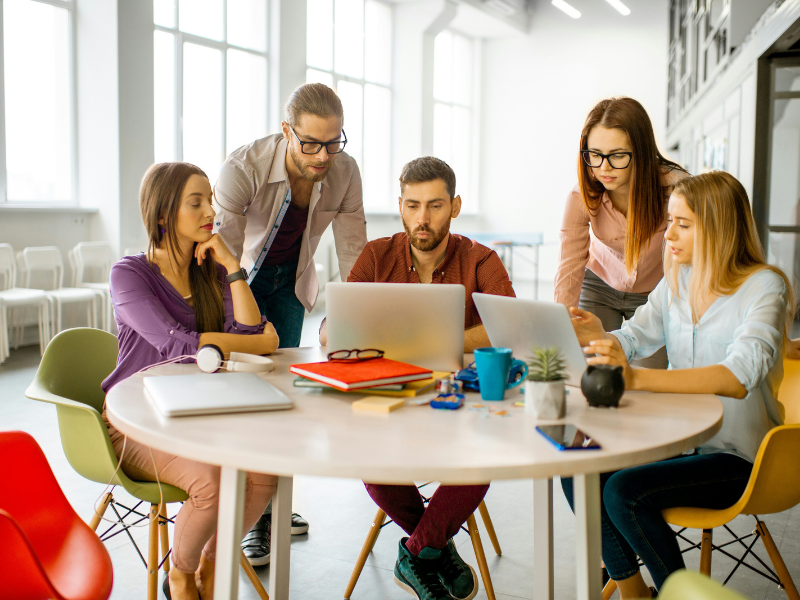 Grupo de jovens profissionais reunidos em torno de uma mesa redonda em um escritório moderno, discutindo ideias e colaborando em um projeto no laptop. A imagem representa trabalho em equipe, criatividade e inovação em um ambiente de trabalho inclusivo.