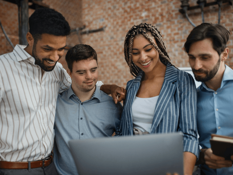 Quatro colegas de trabalho, incluindo uma pessoa com síndrome de Down, estão reunidos em frente a um laptop, sorrindo e colaborando em um projeto. A imagem simboliza inclusão, diversidade e trabalho em equipe em um ambiente corporativo acolhedor