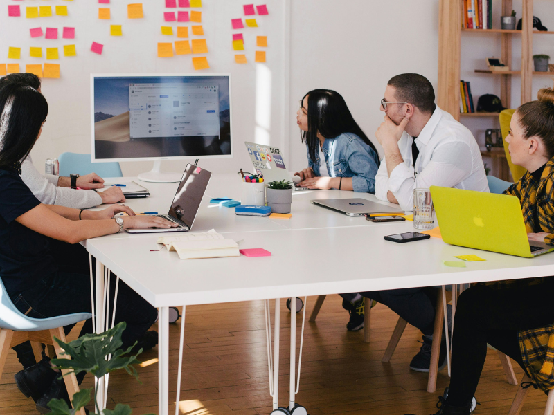 Equipe de trabalho reunida em uma sala de reuniões, concentrada em uma apresentação exibida em um monitor. A mesa está cheia de laptops e materiais de trabalho, enquanto post-its coloridos decoram a parede ao fundo. A imagem transmite colaboração, planejamento e foco no ambiente corporativo.