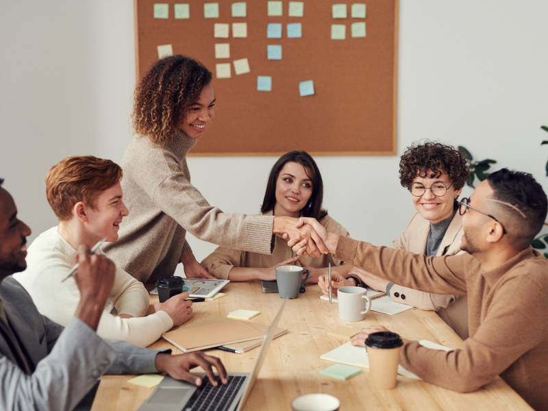 Um grupo diverso de profissionais está reunido em uma mesa de escritório, engajados em uma conversa amigável. No centro da imagem, uma mulher de suéter bege e cabelo cacheado sorri enquanto aperta a mão de um colega, criando um ambiente acolhedor e colaborativo. Os demais participantes observam e interagem, com canecas de café, cadernos e um laptop sobre a mesa. Ao fundo, um quadro de cortiça com notas adesivas sugere planejamento e organização. A cena transmite inclusão, trabalho em equipe e um ambiente corporativo positivo.