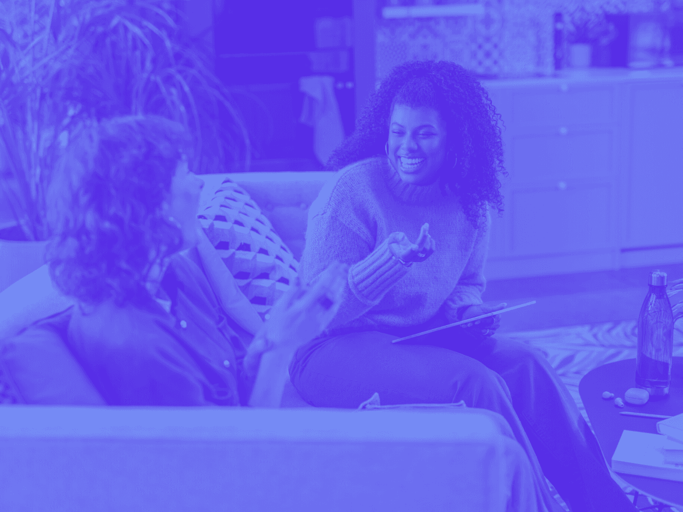 Duas mulheres sentadas em um sofá, conversando sorridentes. Uma delas segura um tablet enquanto parecem estar em uma reunião descontraída.